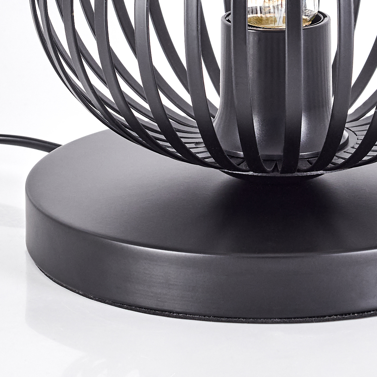 Nahaufnahme einer schwarzen Metallkäfiglampe. Sie hat einen runden Sockel. Der Käfig umgibt eine Glühbirnenfassung. Ein schwarzes Kabel geht vom Sockel aus.