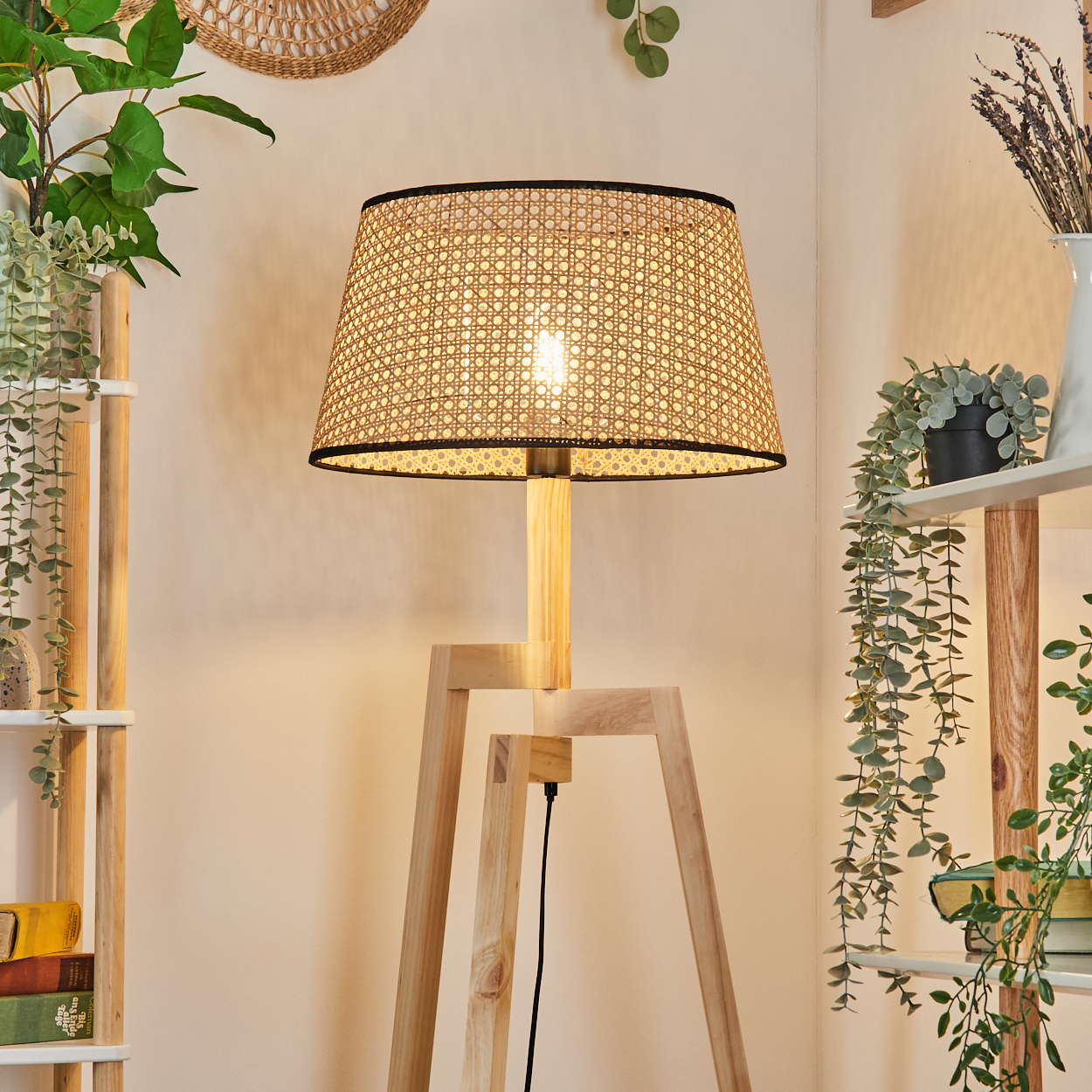 Eine Holz-Stehlampe mit gewebtem Lampenschirm ist beleuchtet. Pflanzen stehen auf Regalen und hängen herunter. Eine gewebte Wanddekoration ist sichtbar.