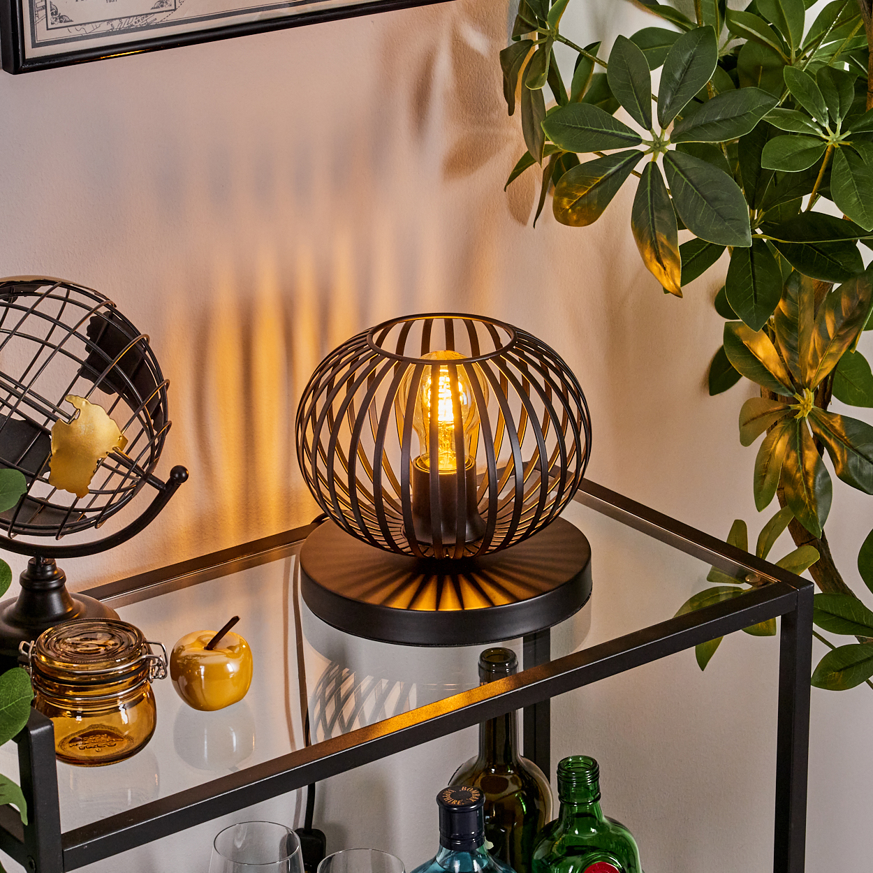 Eine schwarze Metalllampe mit einer leuchtenden Glühbirne steht auf einem Glasregal. Eine kleine Weltkugel, ein Glas und ein Apfel liegen auf dem Regal. Grüne Pflanzen.