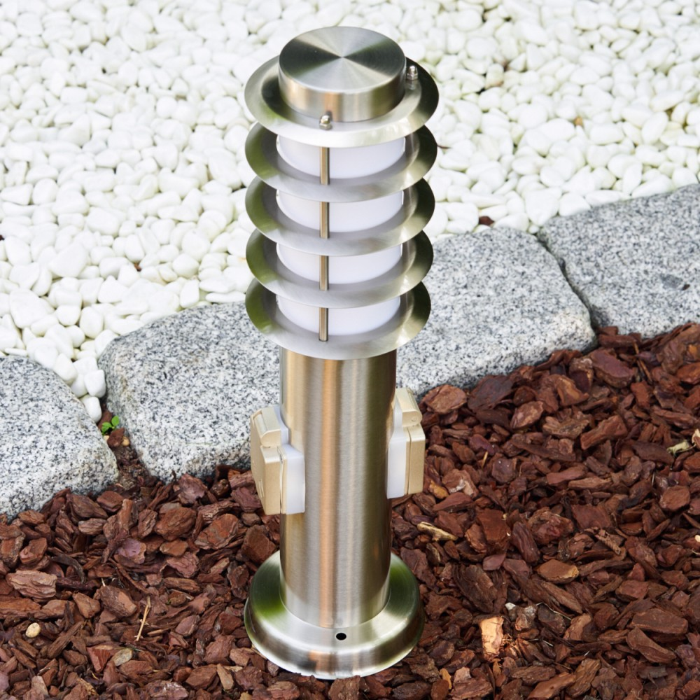 Eine Edelstahl-Außenlampe steht auf braunem Mulch, begrenzt von grauen Steinen und weißen Kieselsteinen. Die Lampe hat ein mehrschichtiges Design.