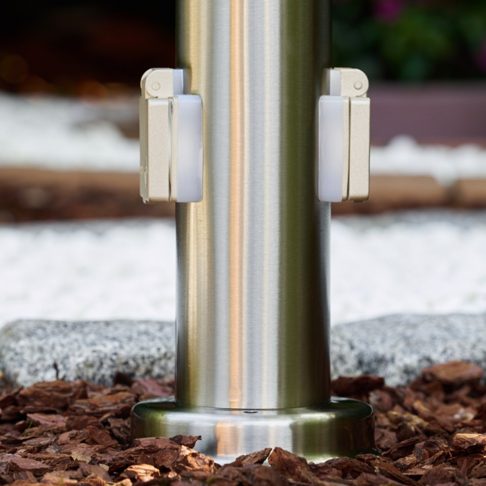 Edelstahlpfosten mit zwei beige-weißen Lichtern. Die Basis steht auf braunem Mulch. Hintergrund hat weiße Steine.