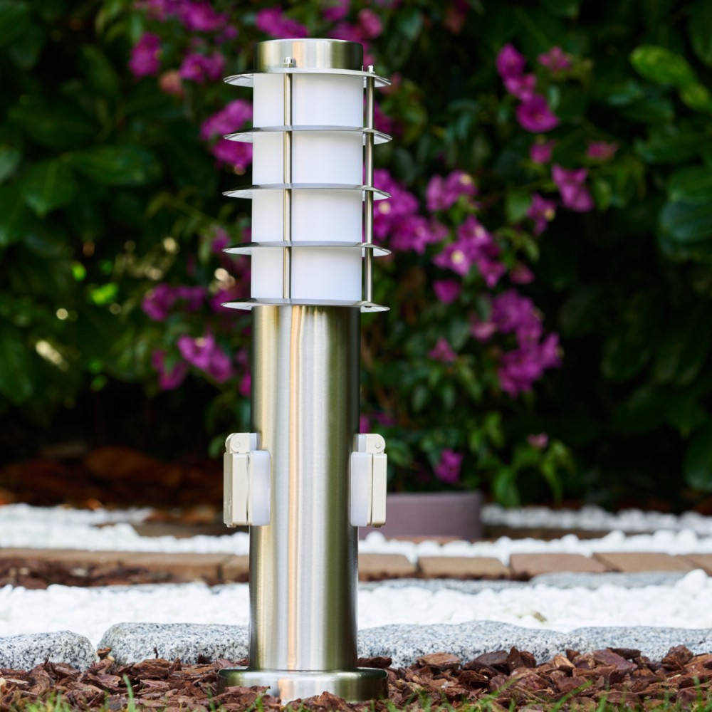 Eine Außenlampe aus Edelstahl steht in einem Garten. Lila Blüten und grünes Laub bilden den Hintergrund. Weißer Kies und Mulch sind sichtbar.