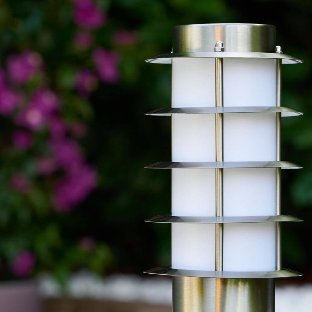 Eine moderne Außenlampe mit zylindrischem weißem Lichtteil und Metallbesatz. Hintergrund ist verschwommenes grünes Laub und rosa Blüten.