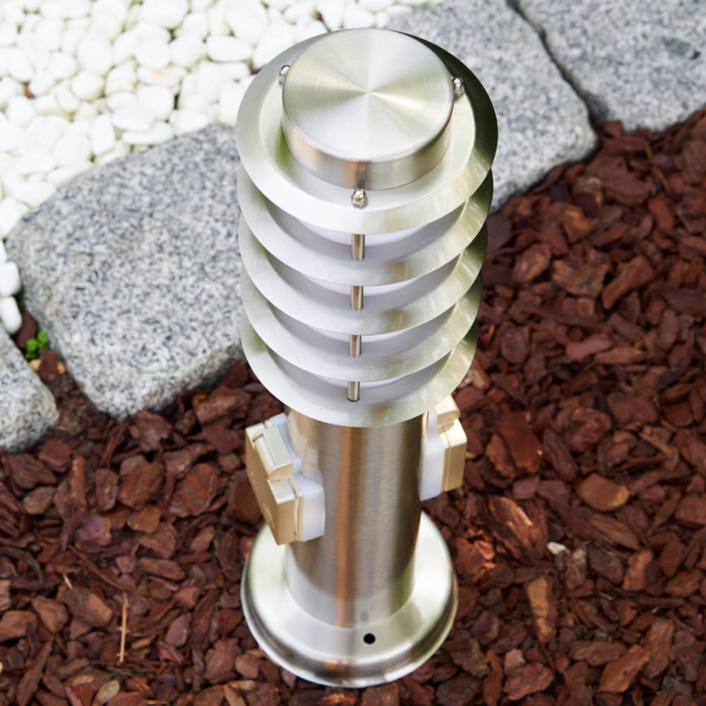 Eine silberne Außenlampe mit kreisförmigem Design steht auf Holzschnitzeln. Steine und weiße Kiesel im Hintergrund.