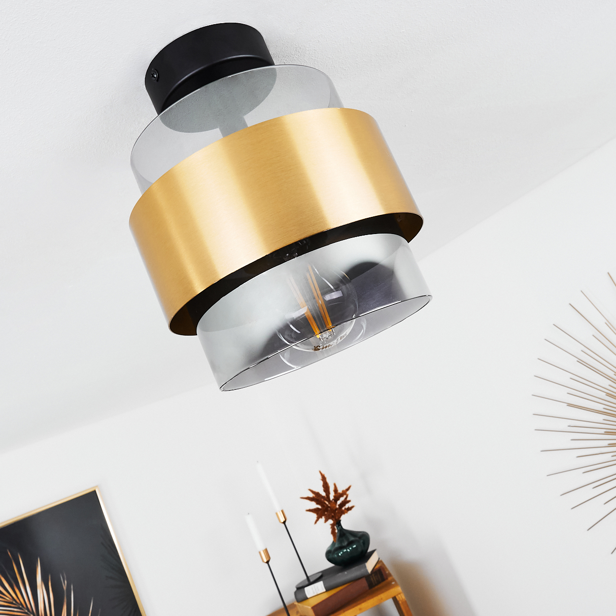 Eine Deckenlampe mit schwarzem Sockel, goldenem Ring und Glasschirm. Darunter ein Regal mit Kerzen, Büchern und einer Vase.