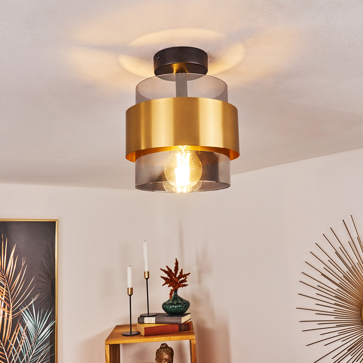 Eine moderne Deckenlampe mit goldenem Band und sichtbarer Glühbirne. Darunter ein Holzregal mit Dekoration und Kerzen.