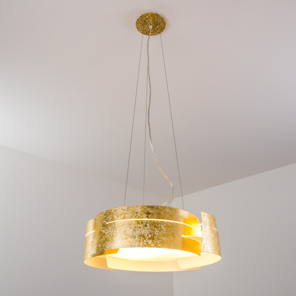Eine Pendelleuchte mit Blattgold hängt von der Decke. Die Lampe hat einen zylindrischen Schirm mit warmem Licht. Der Hintergrund ist weiß.