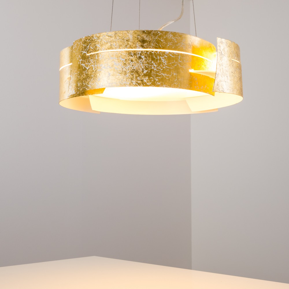 Eine Pendelleuchte aus Blattgold hängt von der Decke. Die Lampe ist kreisförmig und hat einen warmen Schein. Der Hintergrund ist eine hellfarbige Wand.