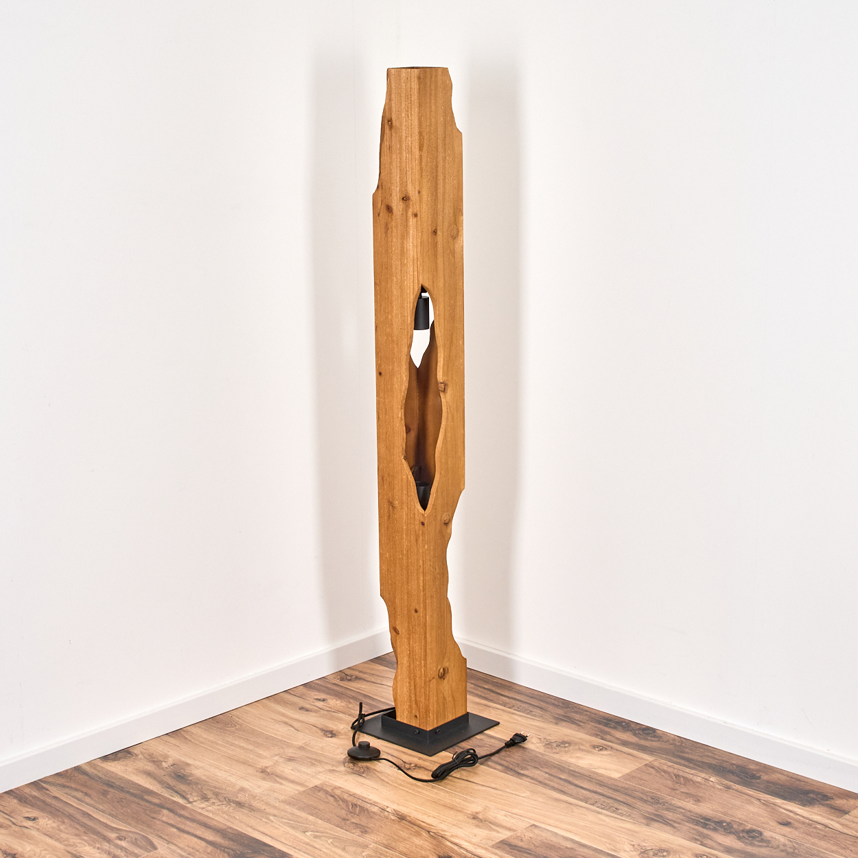 Eine Stehlampe aus Holz mit natürlicher Kante. Sie hat einen rechteckigen Sockel und einen zentralen Ausschnitt. Sie steht in einer Ecke mit weißen Wänden.