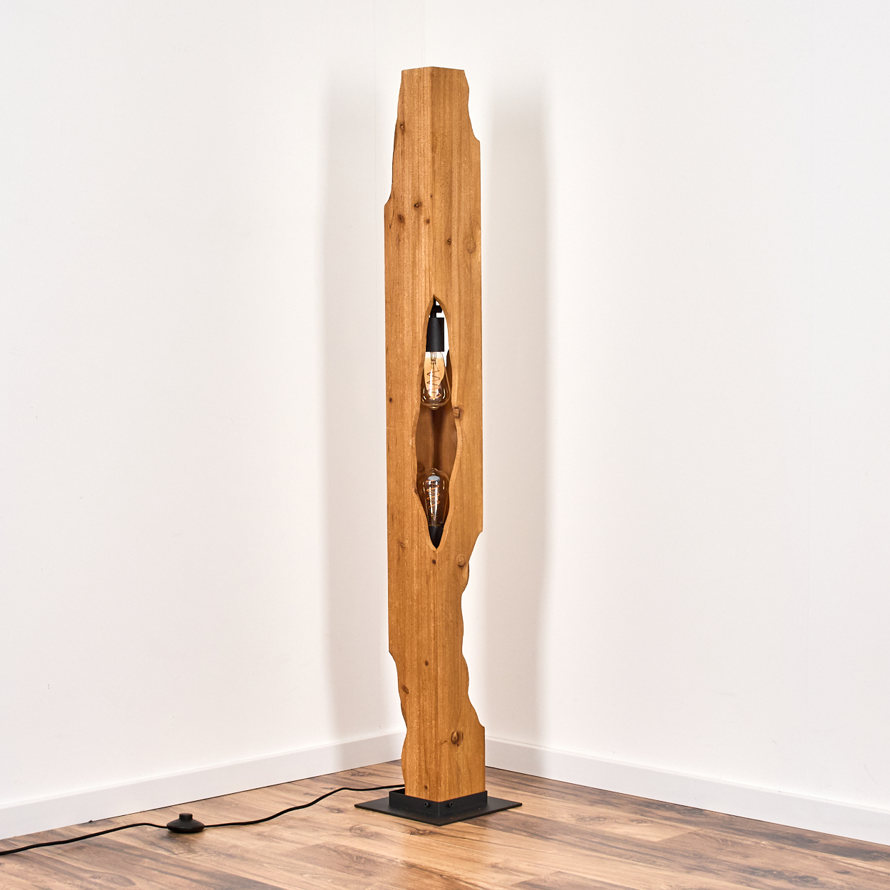 Holzbodenlampe mit zwei freiliegenden Glühbirnen, vor einer weißen Wand. Die Lampe hat einen schwarzen Sockel und ein Netzkabel.