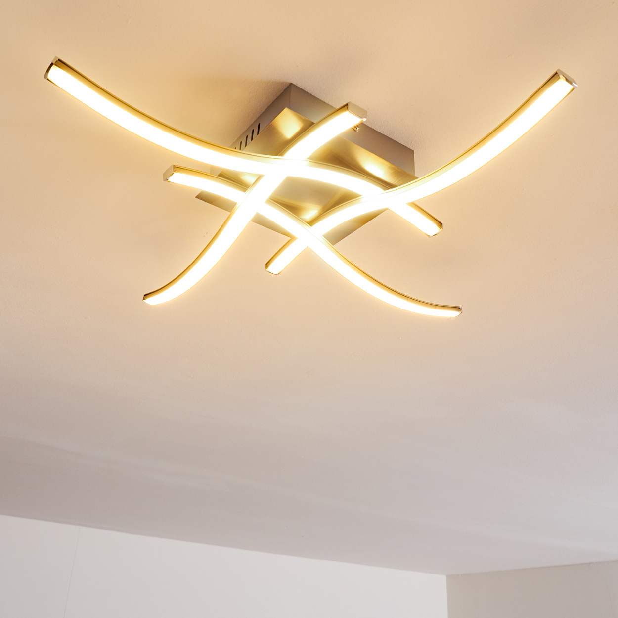 Eine moderne Deckenleuchte mit geschwungenen, leuchtenden weißen Lichtelementen vor einer weißen Decke.