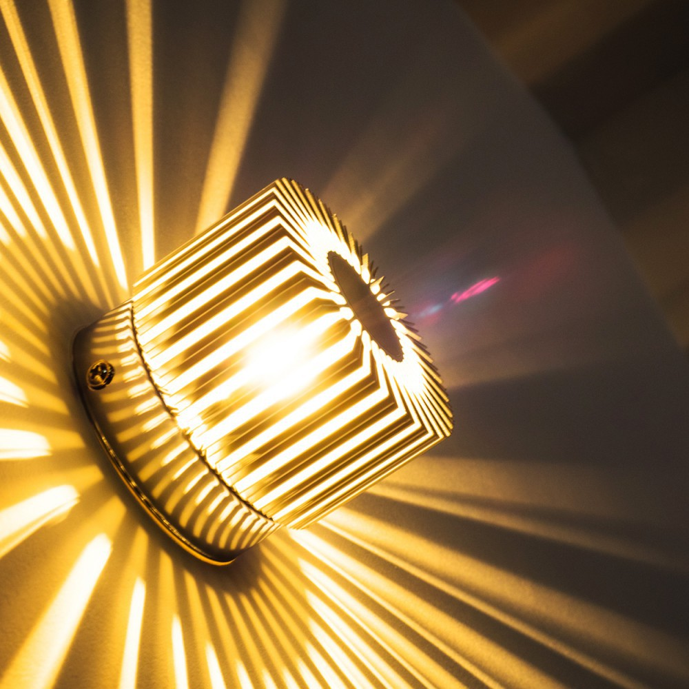 Eine moderne Lampe mit geometrischem Design wirft ein gemustertes Licht auf die Wand. Die Lampe leuchtet und erzeugt eine warme, gelbe Atmosphäre.