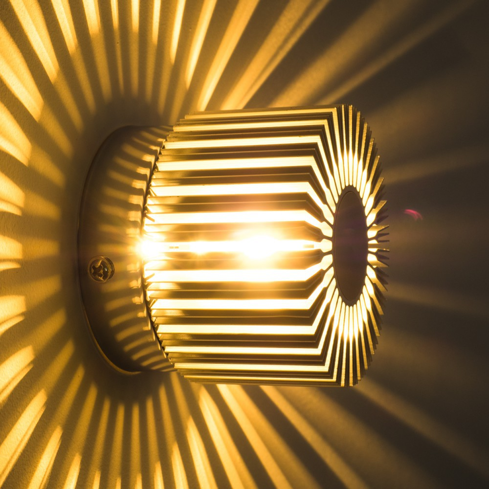 Eine Wandlampe mit zylindrischer Form, die helles Licht aussendet. Ihr Design wirft radiale Schatten auf die Wand. Die Lampe ist metallisch.