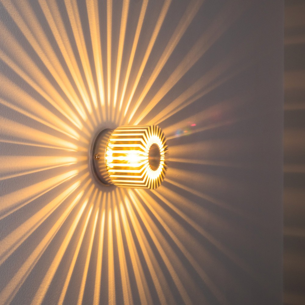Eine Wandleuchte wirft sternförmige Schatten. Die Leuchte hat ein zylindrisches, geometrisches Design und warmes Licht.