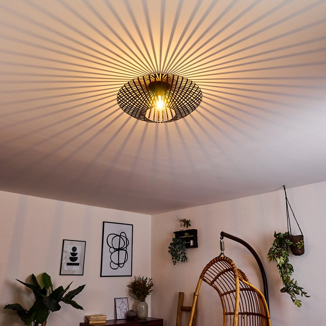 Eine schwarze Deckenlampe wirft radiale Schatten. Der Raum hat weiße Wände, Kunst, Pflanzen und einen Korbschaukelstuhl.