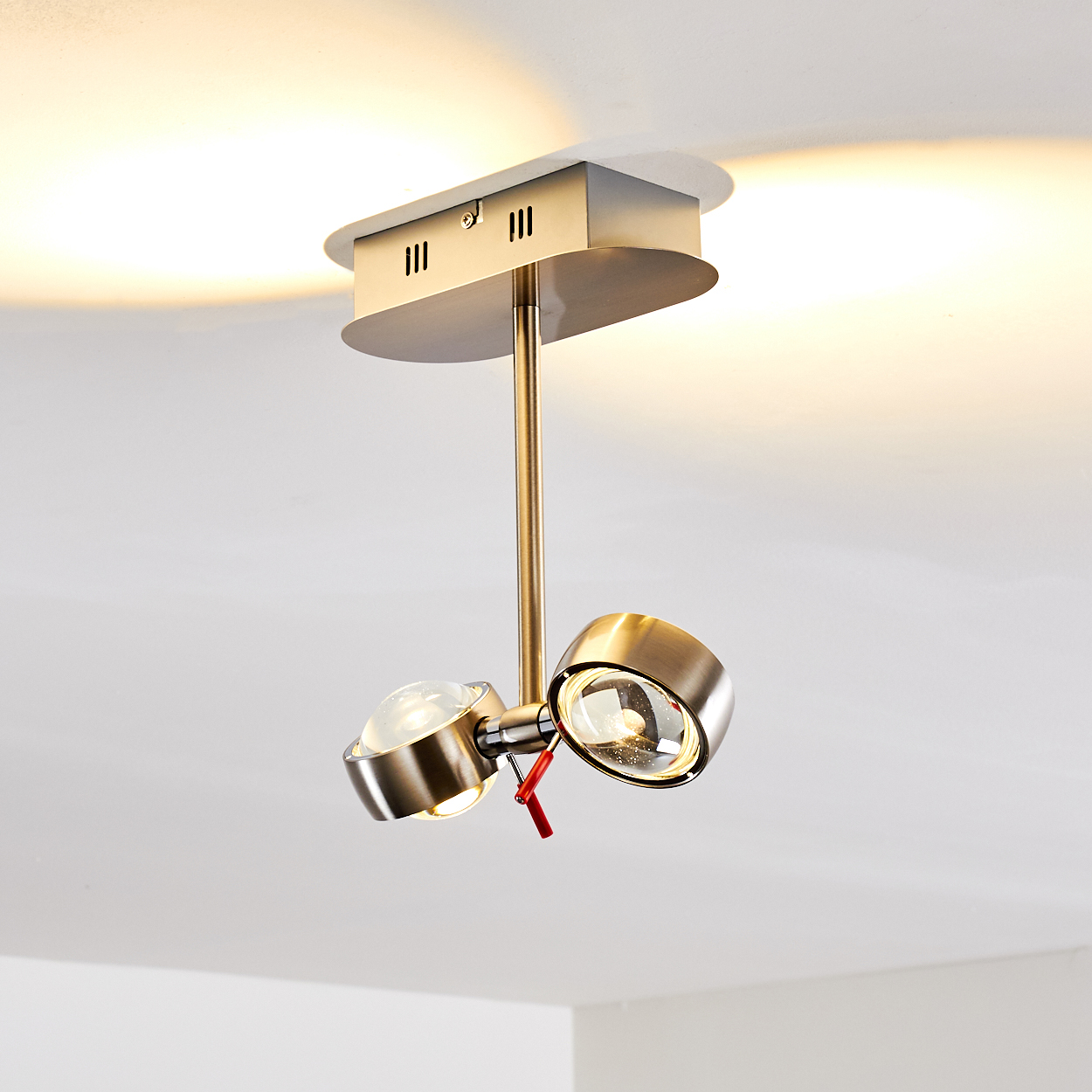 Eine Deckenstrahler-Leuchte mit zwei verstellbaren Lampen. Die Leuchte ist metallisch, mit rotem Verstellhebel. Der Hintergrund ist weiß.