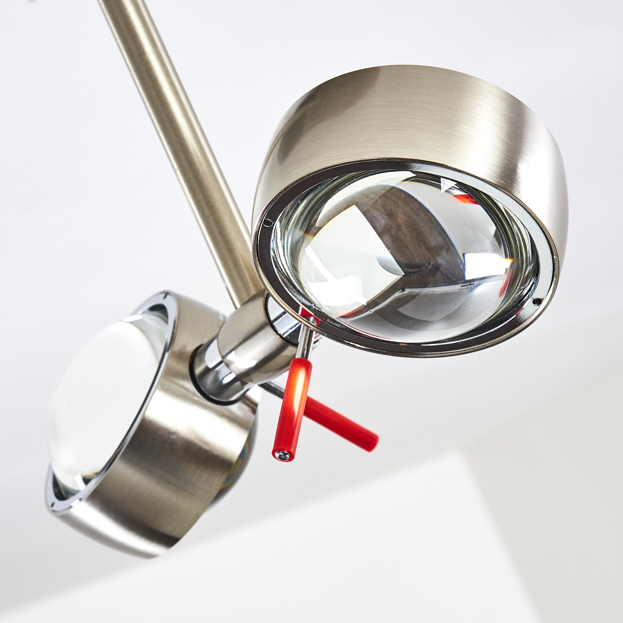 Nahaufnahme einer modernen Metalllampe mit zwei Leuchtköpfen. Jeder Kopf hat eine Glaslinse. Rote Akzente sind sichtbar. Der Hintergrund ist weiß.
