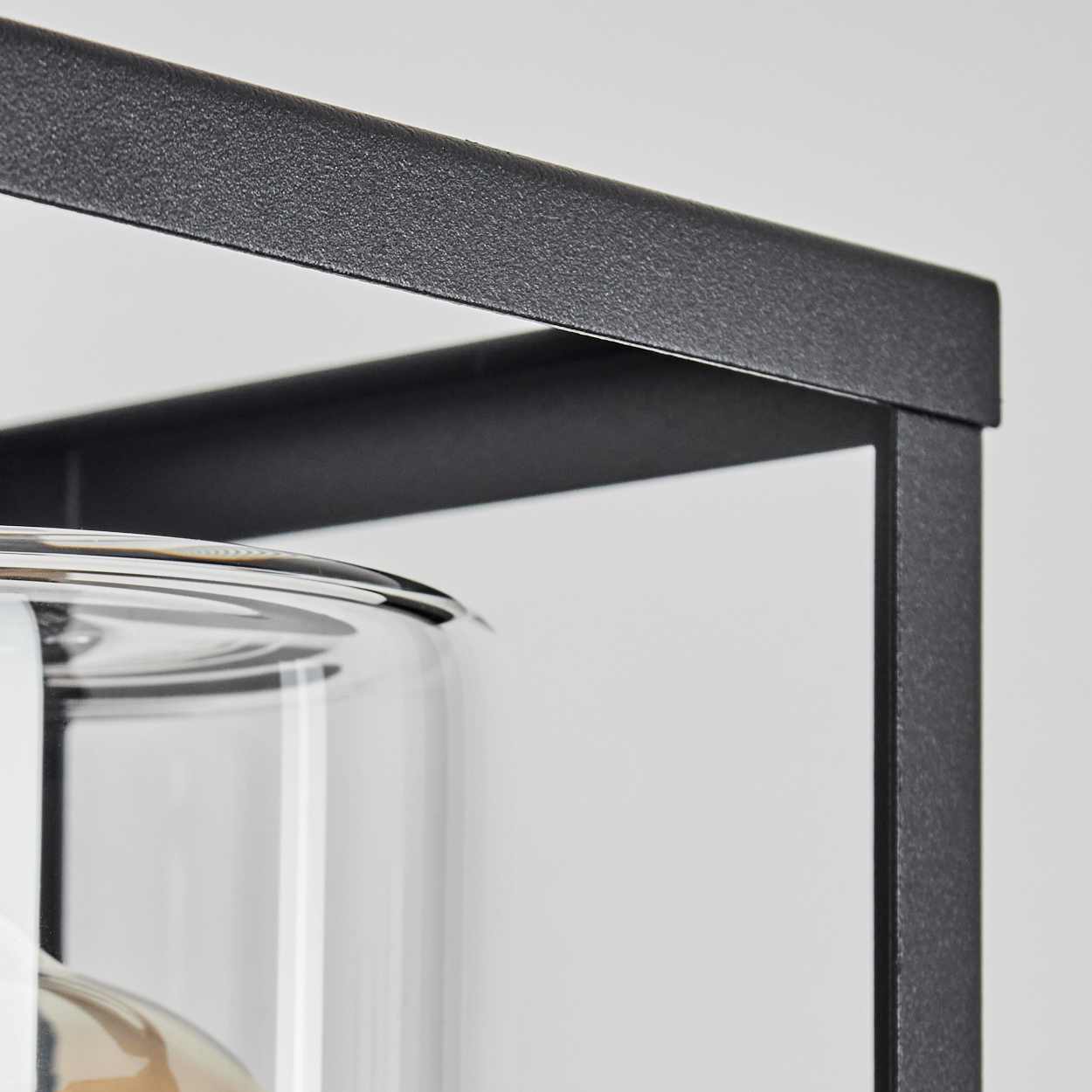 Nahaufnahme eines schwarzen Metallrahmens mit einem durchsichtigen Glaszylinder in der linken unteren Ecke. Der Hintergrund ist schlicht weiß.