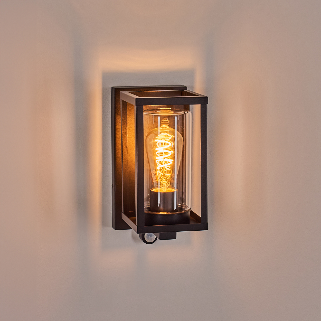 Schwarz-Wandlampe mit Glaszylinder. Die Glühbirne leuchtet. Der Hintergrund ist eine hellbeige Wand mit Schatten.
