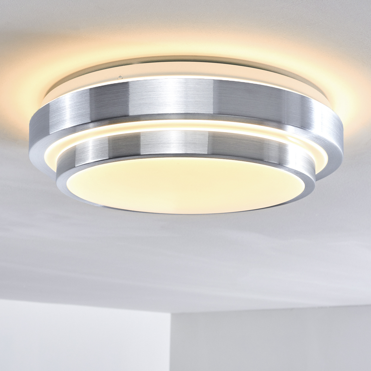 Eine moderne, runde Deckenleuchte mit gebürsteter Metalloberfläche. Sie strahlt ein warmes, diffuses Licht aus, vor einer weißen Decke.