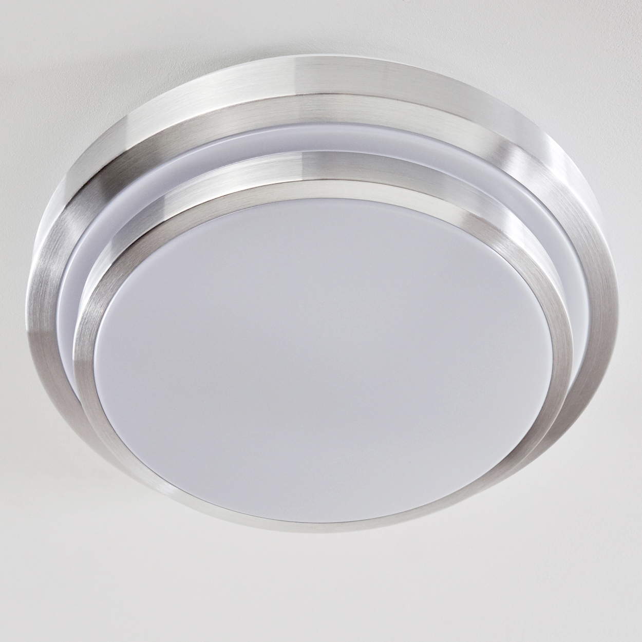 Eine kreisförmige Deckenleuchte mit gebürsteter Metallverkleidung umgibt eine weiße, undurchsichtige Lichtplatte. Der Hintergrund ist eine schlichte weiße Decke.