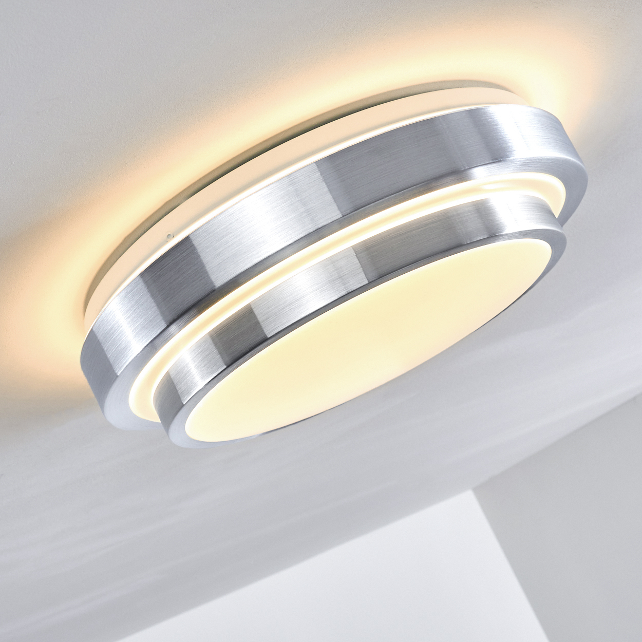 Eine moderne, metallische Deckenleuchte. Sie hat ein kreisförmiges Design mit geschichteten Ringen, die ein warmes, weiches Licht ausstrahlen. Die Decke ist weiß.