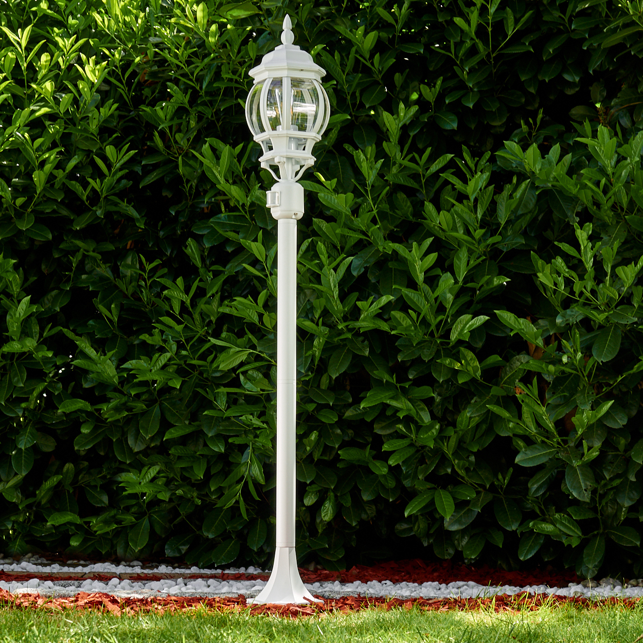 Eine weiße Laterne steht vor einer grünen Hecke. Die Lampe ist beleuchtet. Der Boden hat Gras, weiße Steine und rote Rinde.