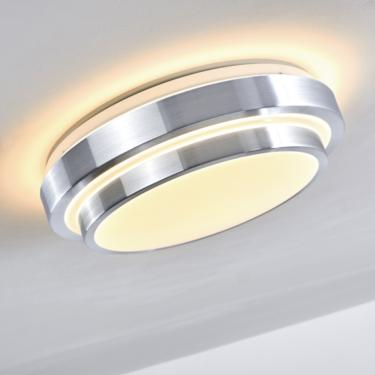 Eine moderne, kreisförmige Deckenleuchte. Sie hat einen silbernen Metallrahmen und strahlt ein weiches, warmes Licht aus. Sie ist an einer weißen Decke montiert.