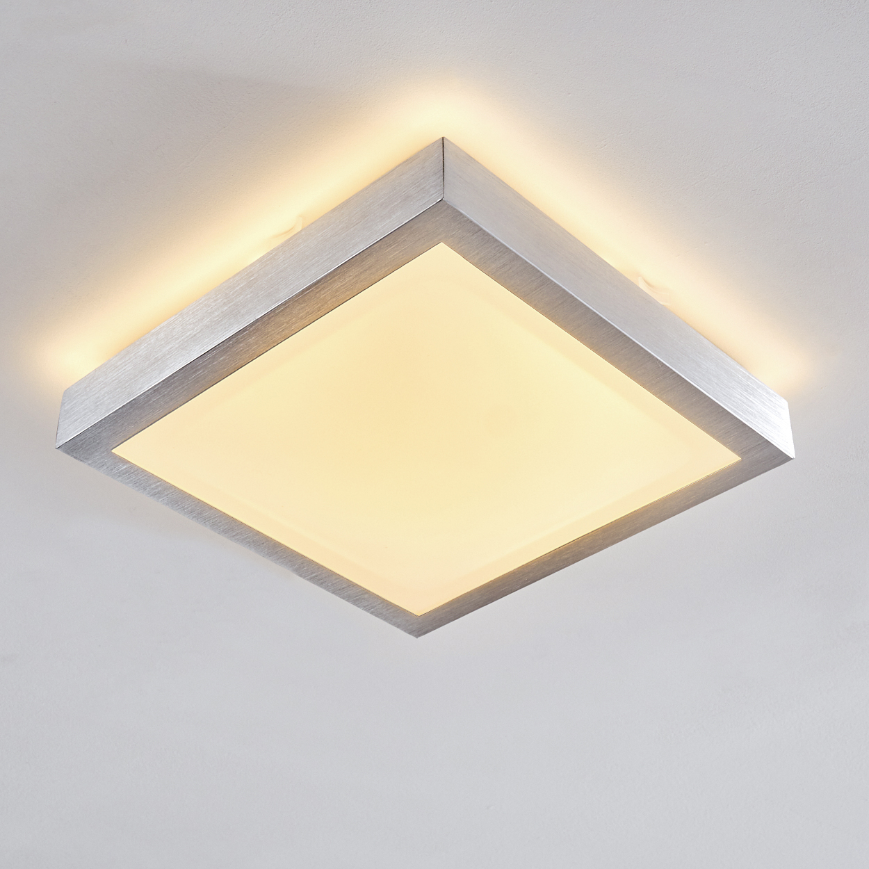 Eine quadratische Deckenleuchte. Sie hat einen silbernen Rahmen und eine weiche, gelbe Lichtfläche. Der Hintergrund ist eine schlichte weiße Decke.