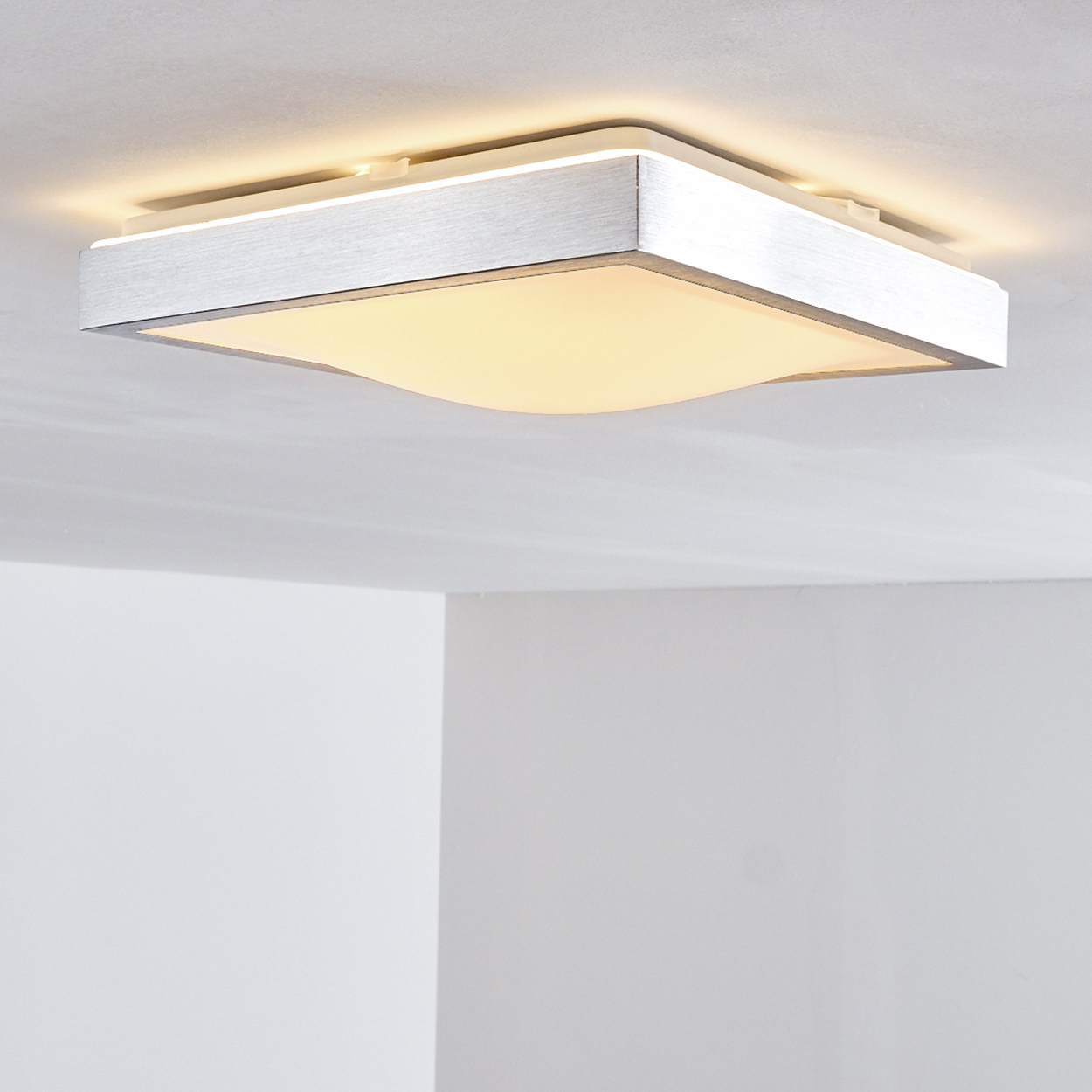 Eine quadratische Deckenlampe aus Metall mit einem weißen, gebogenen Lampenschirm. Die Lampe leuchtet und wirft ein warmes Licht. Weiße Wände sind im Hintergrund.