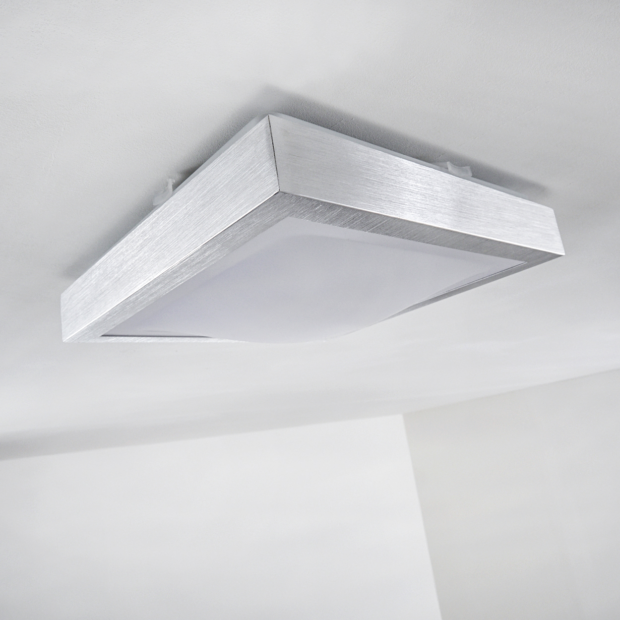 Eine quadratische Deckenleuchte mit silbernem Rahmen. Das Licht hat eine weiße, durchscheinende Platte. Die Decke ist weiß.