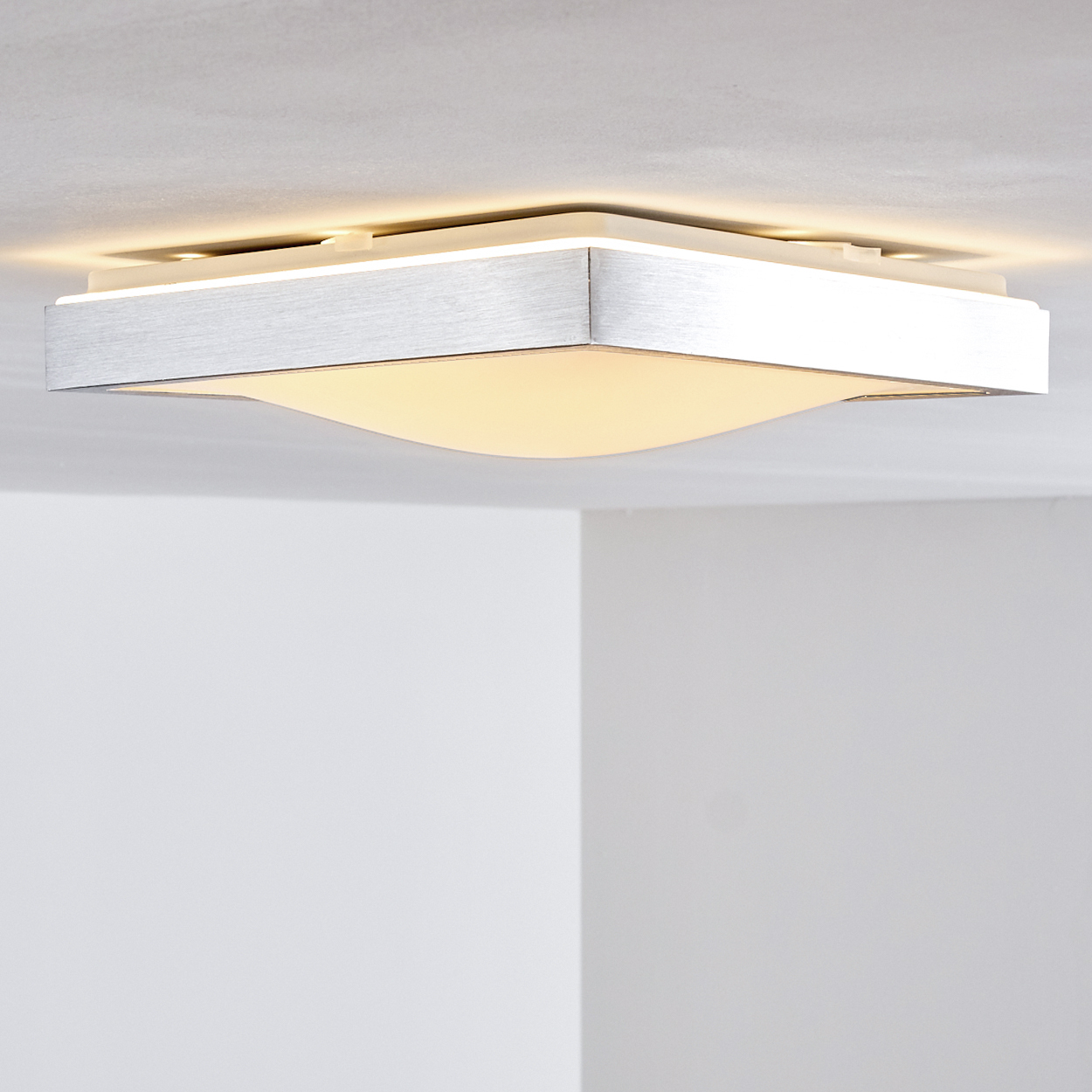 Eine moderne Deckenleuchte mit silberner Verkleidung und einem geschwungenen, leuchtenden weißen Diffusor. Weiße Wände sind im Hintergrund.