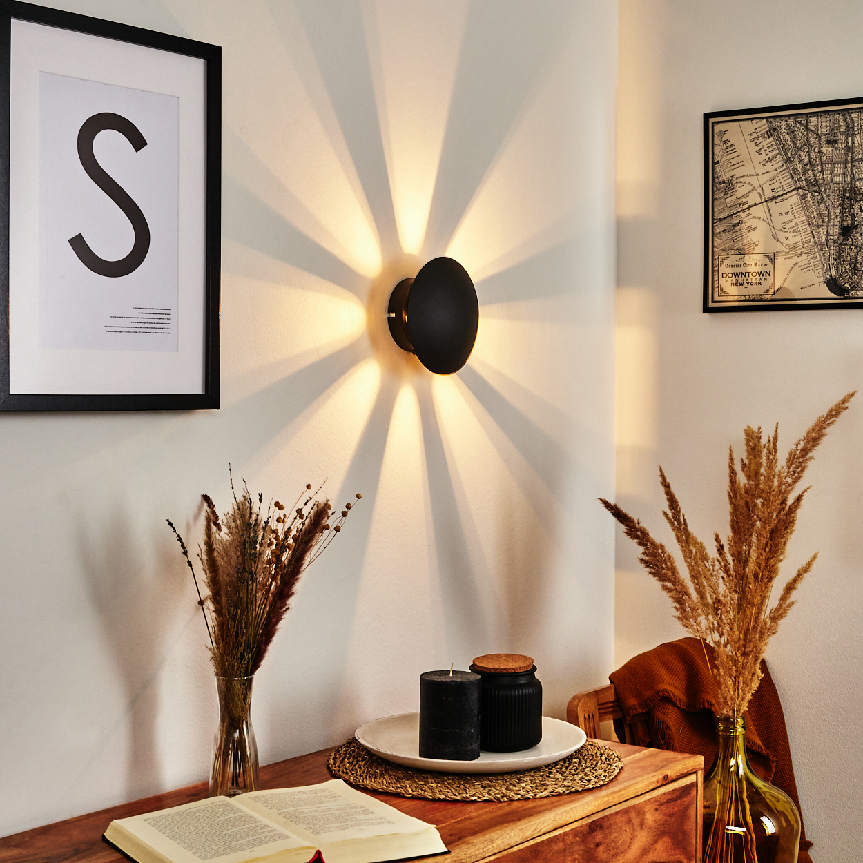 Eine schwarze Wandleuchte wirft Lichtstrahlen. Ein gerahmtes 'S'-Kunstwerk, Trockenblumen, Kerzen, Kartenkunst und ein offenes Buch schmücken eine Holzoberfläche.