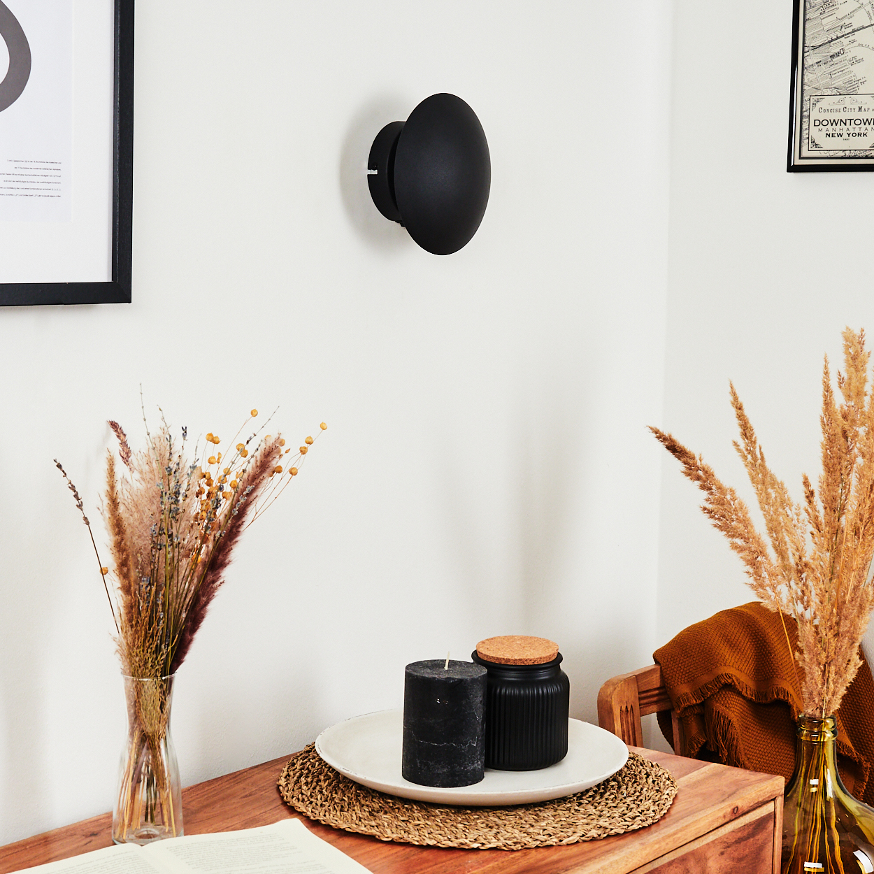 Eine schwarze Wandleuchte, Kerzen und eine Vase mit getrockneten Blumen auf einem Holztisch. Ein gerahmtes Kunstwerk hängt an der Wand.
