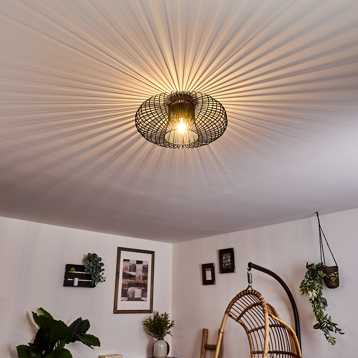 Eine schwarze Deckenlampe aus Draht wirft strahlenförmige Schatten. Darunter ein Raum mit Kunst, Hängepflanzen und einem Rattanstuhl.
