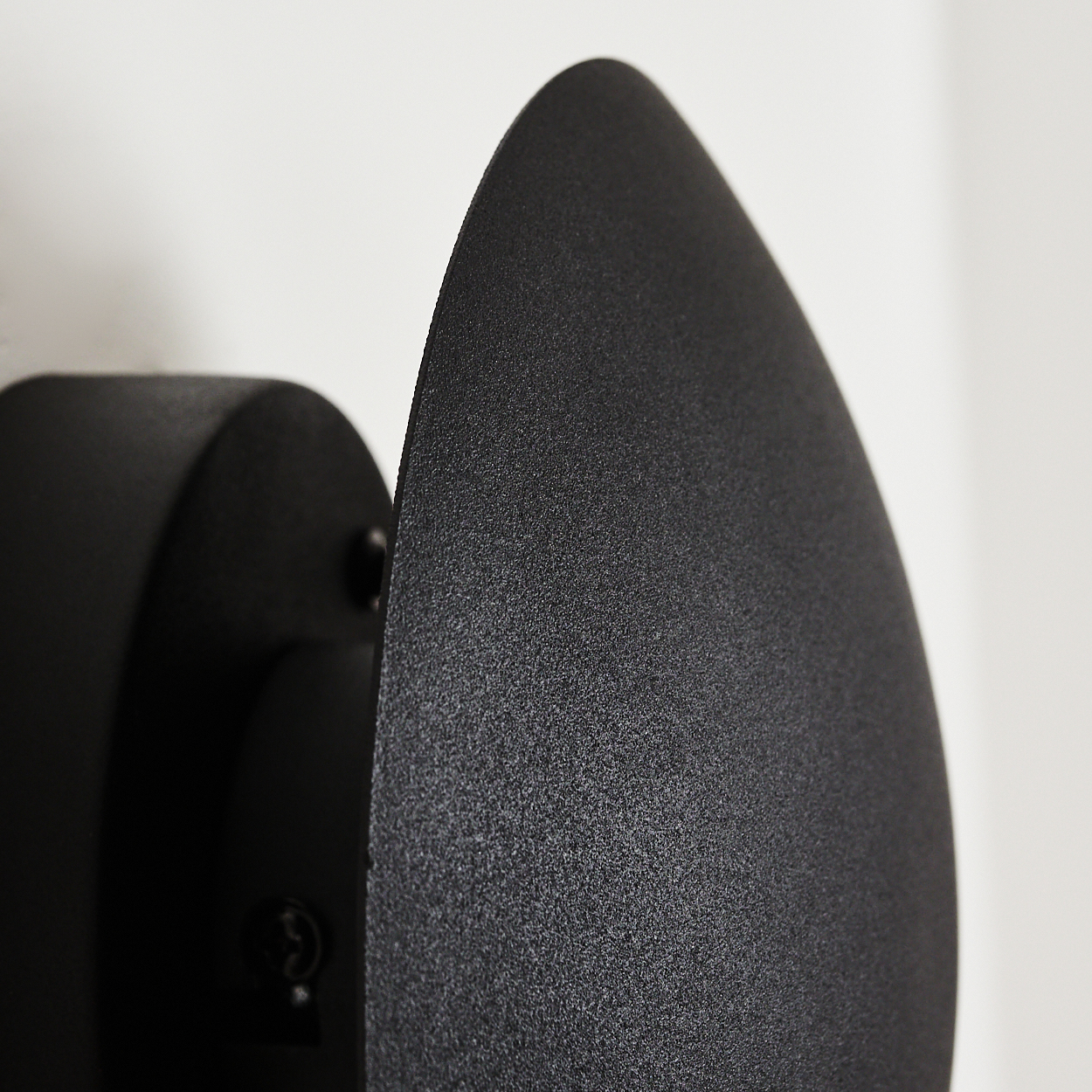Nahaufnahme einer schwarzen, tropfenförmigen Wandleuchte. Die Lampe hat eine strukturierte Oberfläche und ist an einer weißen Wand montiert.