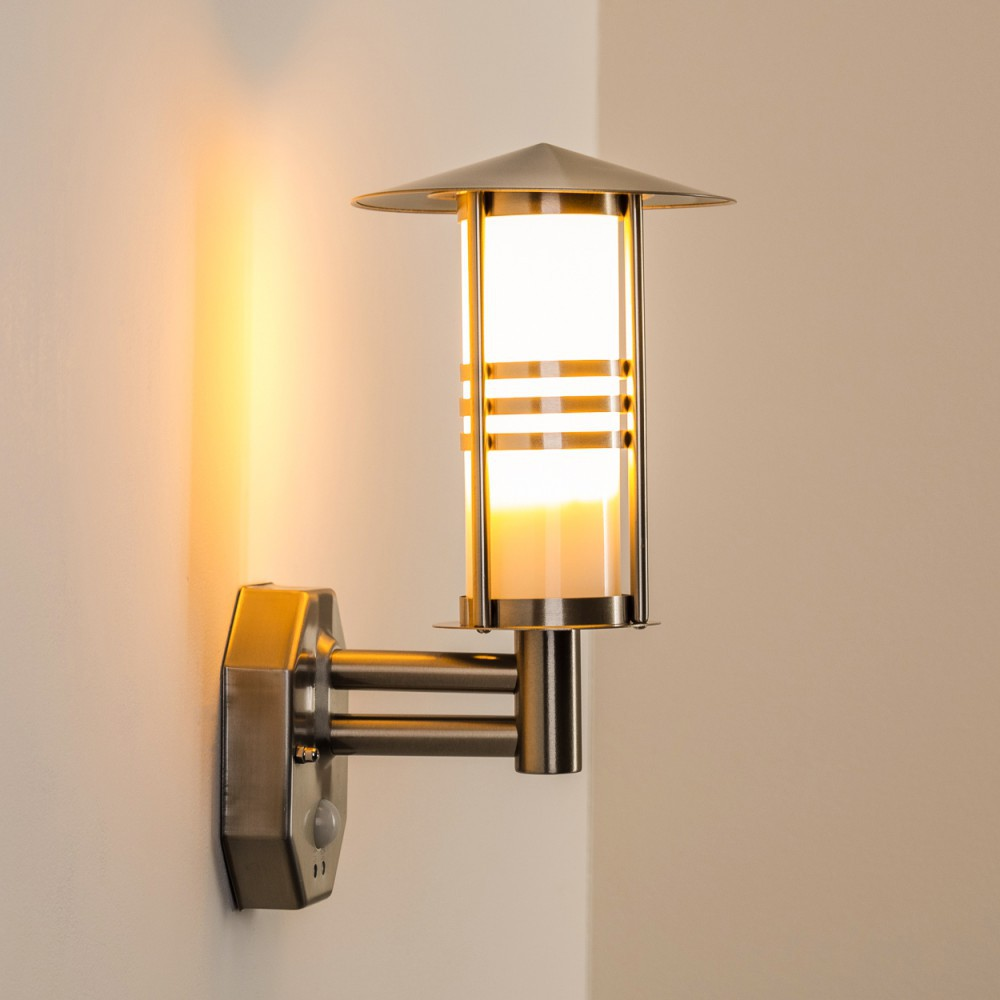 Eine Wandleuchte im Freien mit silbernem Finish leuchtet und wirft einen warmen Schein. Die Lampe hat einen zylindrischen Glasschirm und einen Sensor.