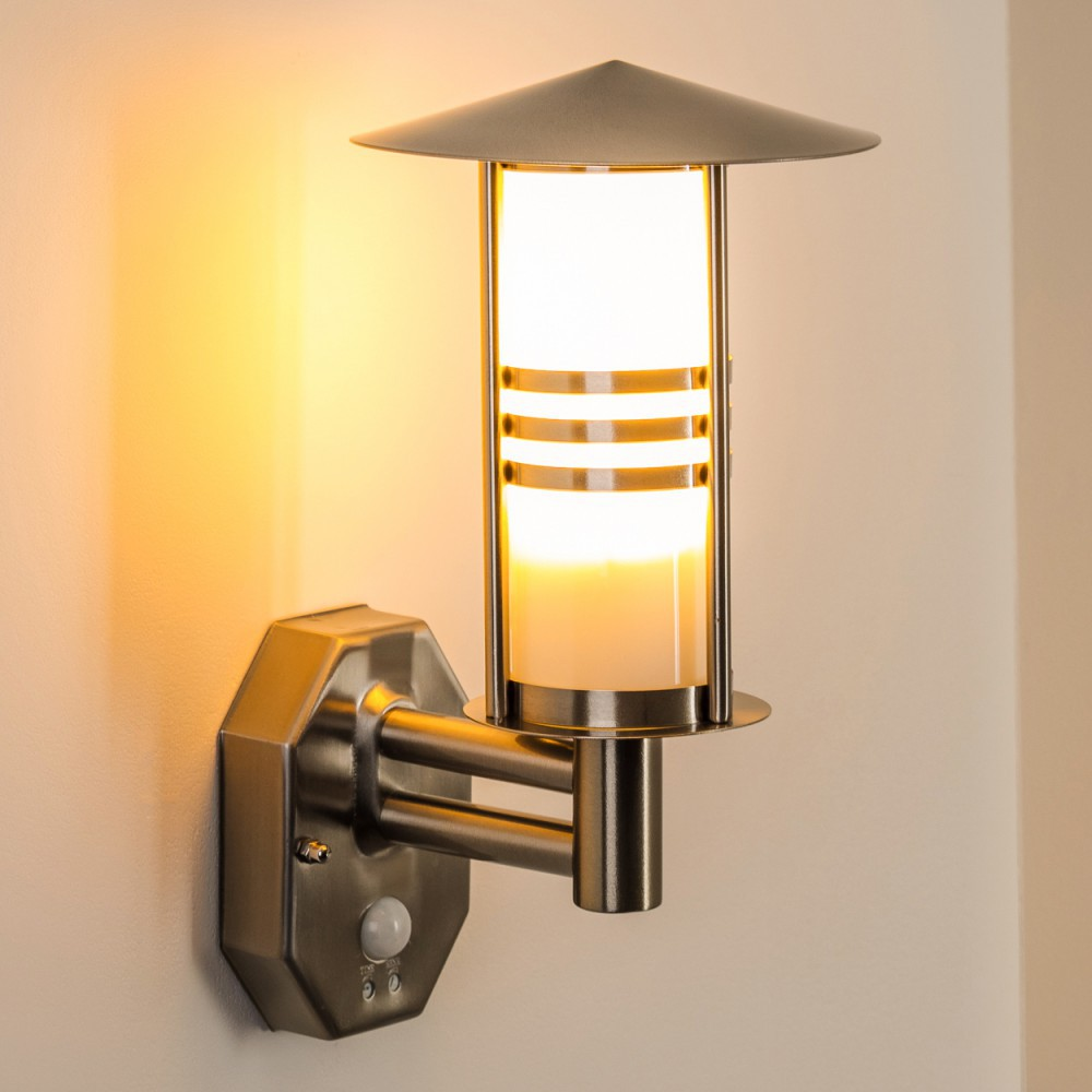 Eine an der Wand montierte Außenlampe mit einem beleuchteten zylindrischen Schirm. Sie hat eine graue Metalloberfläche und ist an einer weißen Wand befestigt.