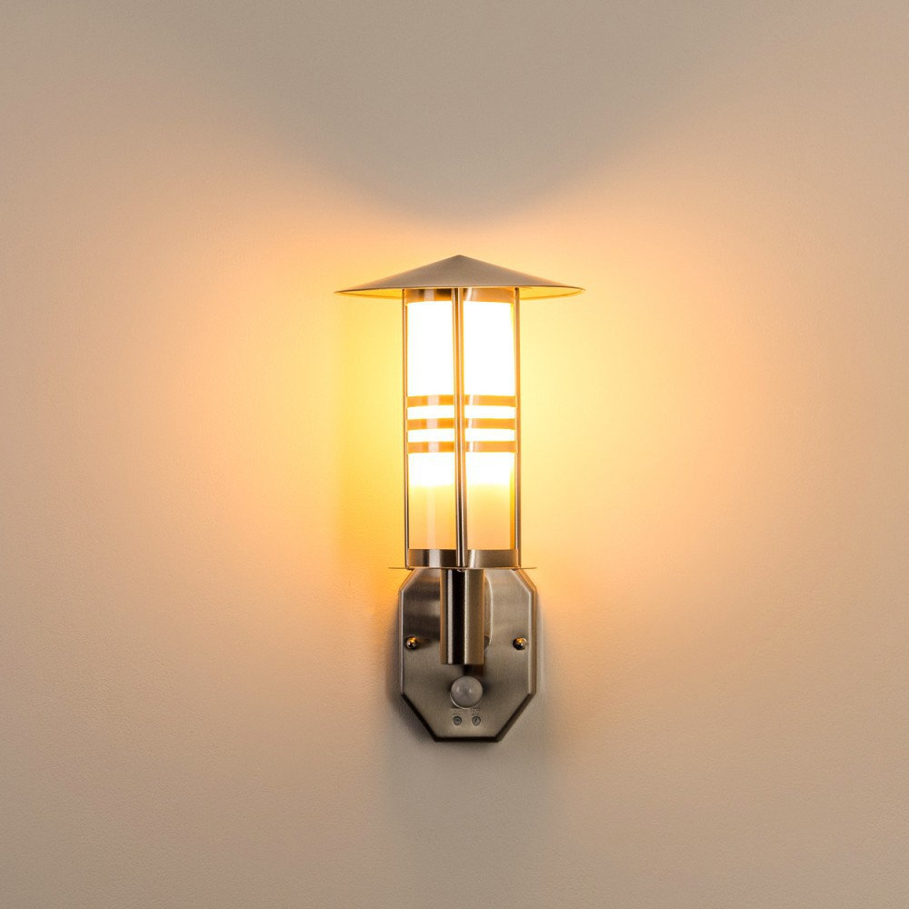 Eine Wand-Außenlampe mit silbernem Finish und Bewegungssensor, die eine Wand warm beleuchtet.