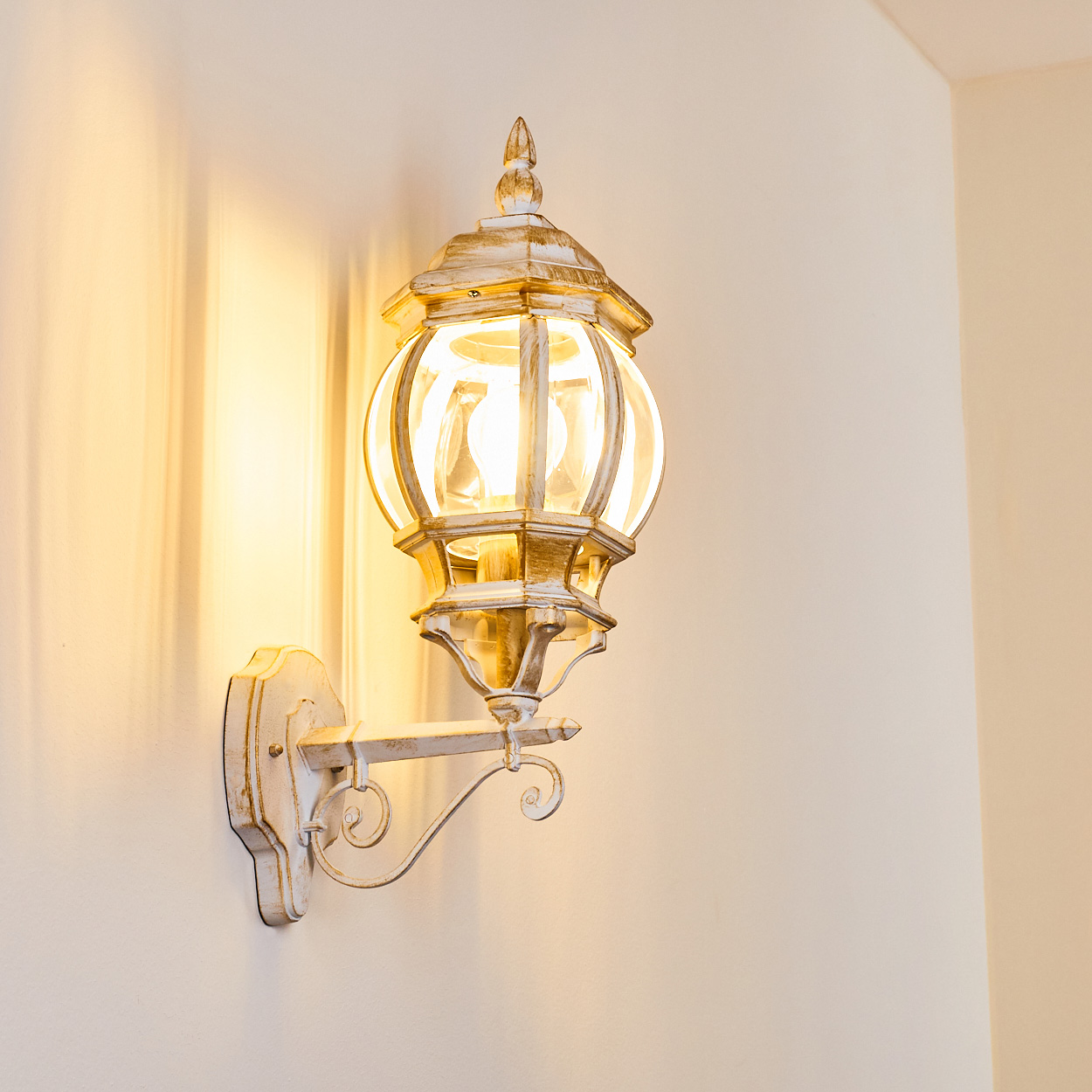 Eine an der Wand befestigte, verzierte, weiß-goldene Laterne mit einer leuchtenden Glühbirne. Die Laterne wirft einen warmen Schein auf die cremefarbene Wand.