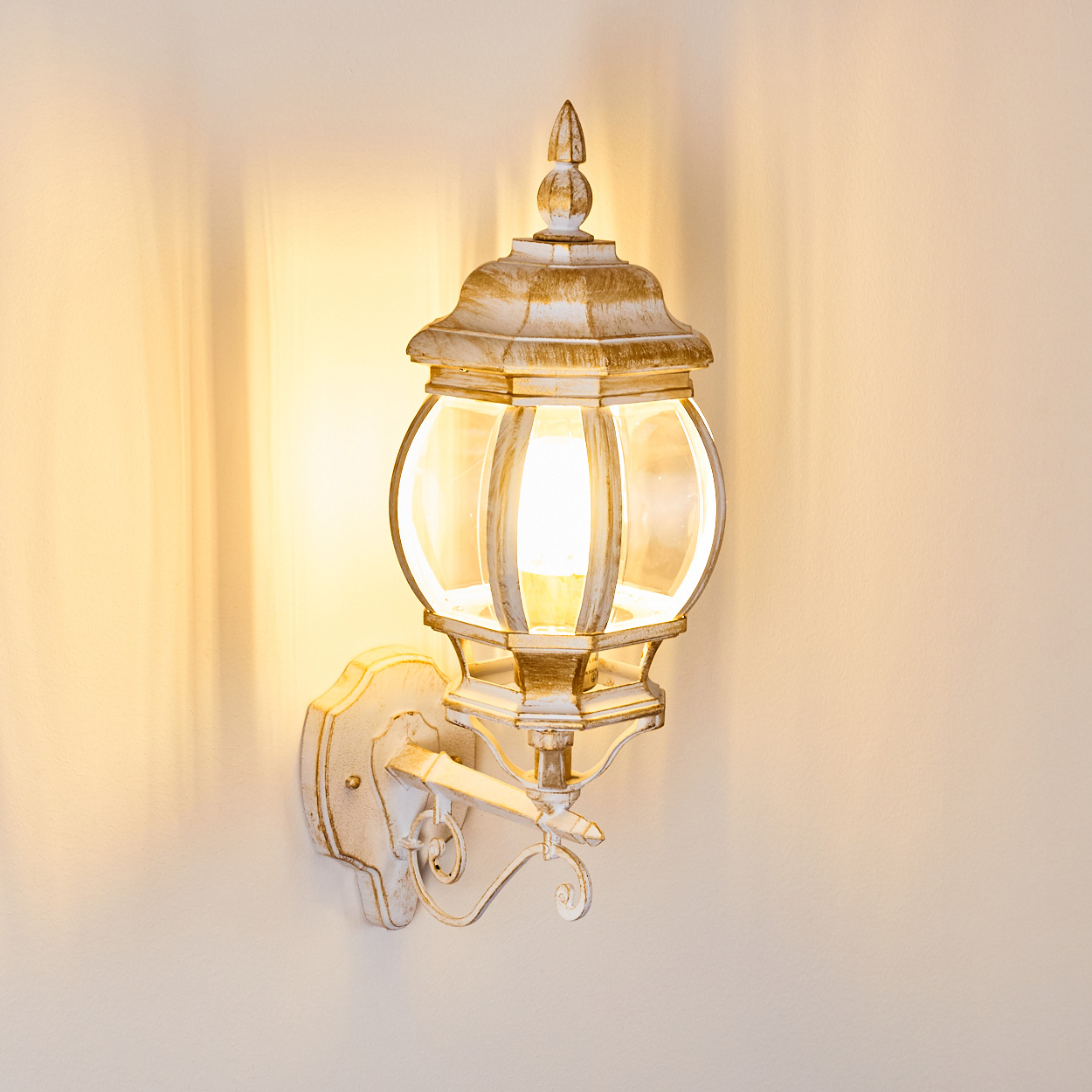 Eine Wandleuchte für den Außenbereich mit weiß-goldener Oberfläche. Sie hat eine Glaskugel und wirft Licht auf die Wand.