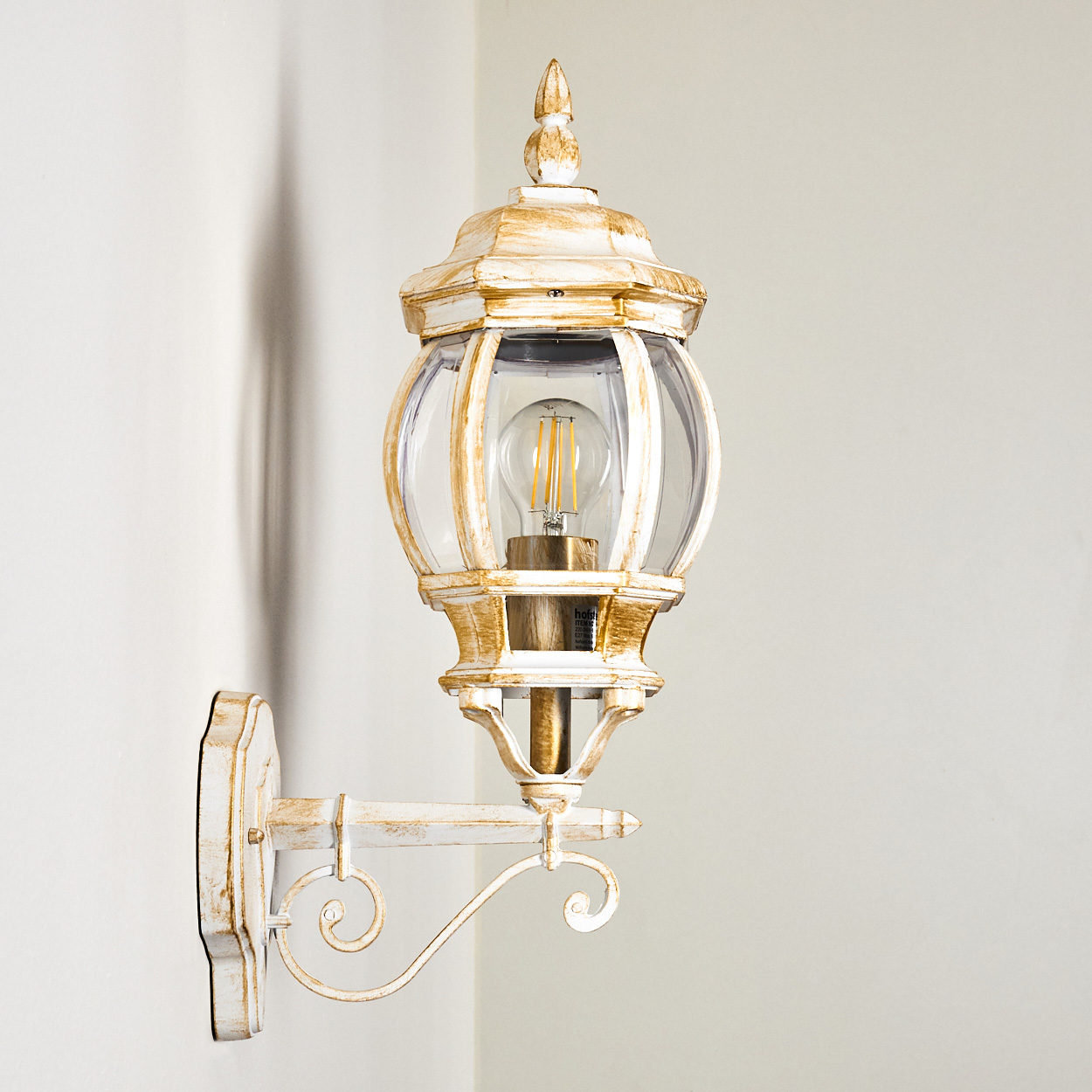 Eine weiß-goldene Außenlampe an einer weißen Wand. Die Lampe hat ein klares Glasgehäuse mit einer sichtbaren Glühbirne.