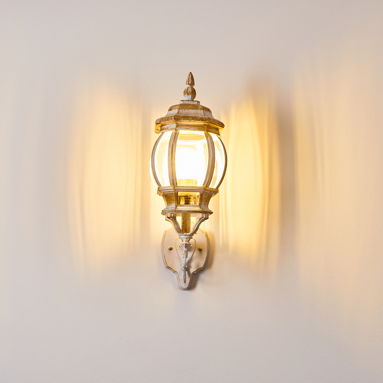 Eine antike weiße Wandlampe mit leuchtender Glühbirne ist an einer hellbeigen Wand angebracht. Licht reflektiert von der Lampe.
