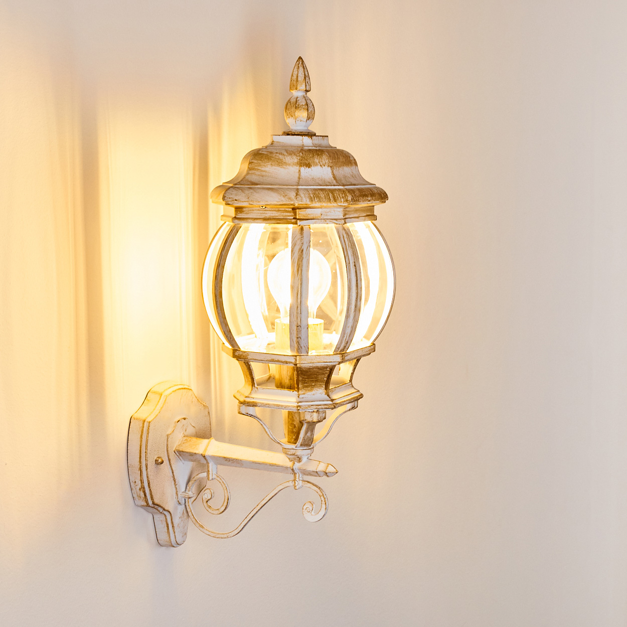 Eine verzierte, weiß-goldene Wandlampe mit Glaskugel. Sie leuchtet und ist an einer weißen Wand befestigt und wirft einen warmen Schein.