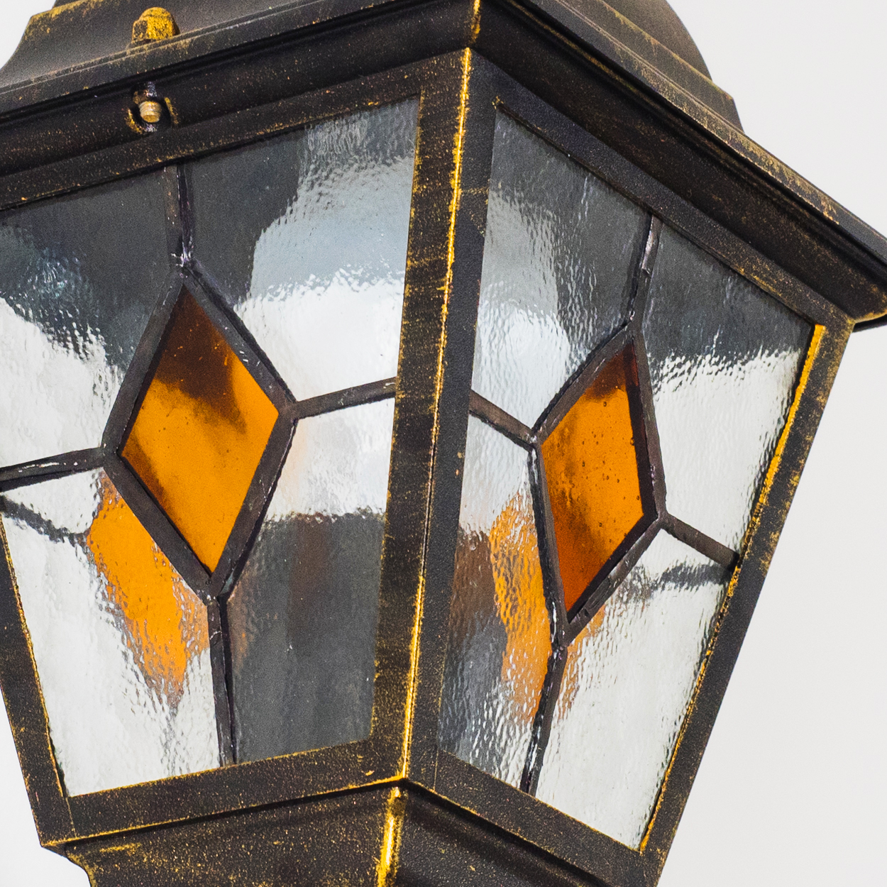 Nahaufnahme einer verzierten schwarz-goldenen Lampe. Sie hat Glasscheiben mit rautenförmigen bernsteinfarbenen Akzenten vor weißem Hintergrund.