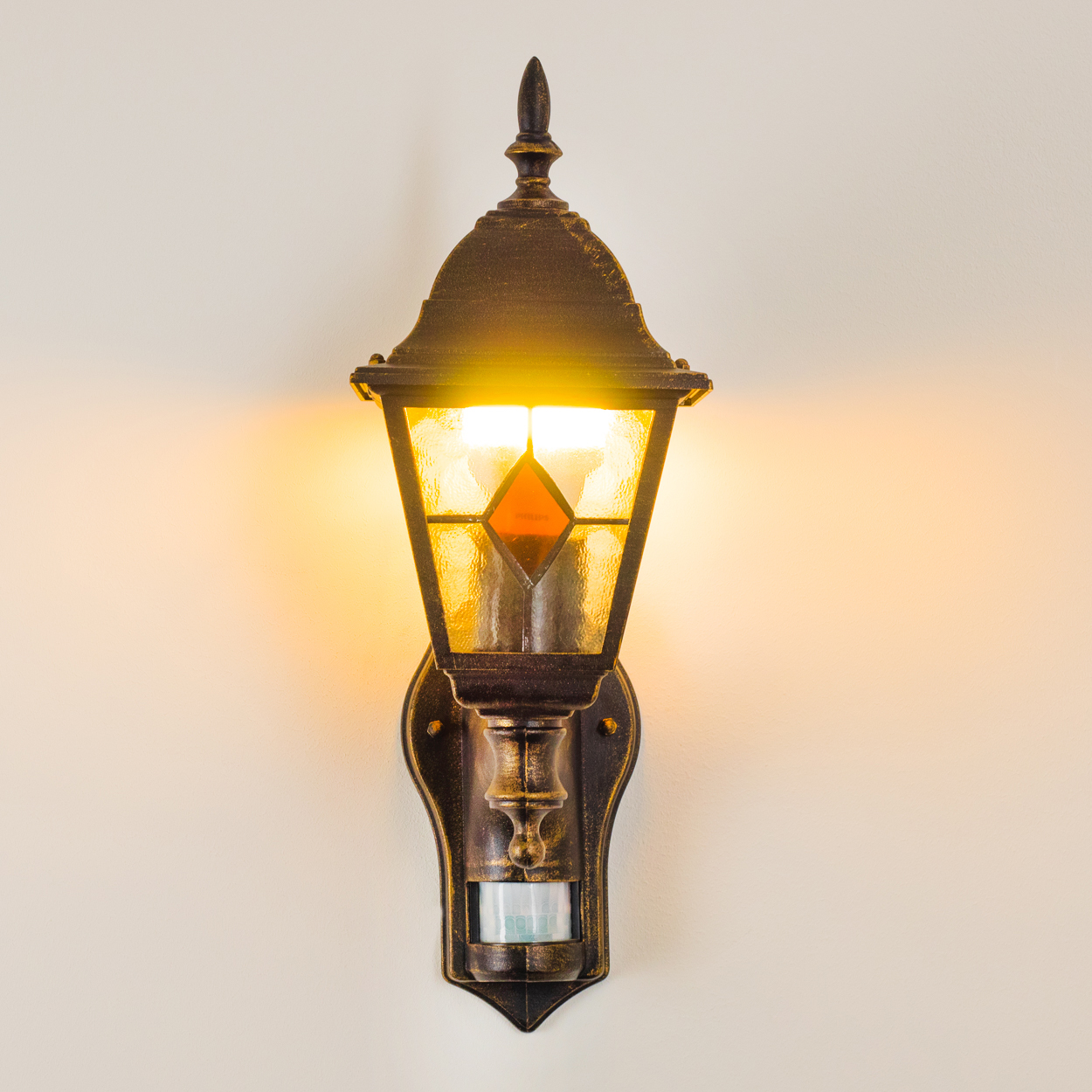 Eine verzierte, bronzefarbene Außenlampe mit einem beleuchteten Lampenschirm im Glasmalerei-Stil ist an einer cremefarbenen Wand befestigt. Unten befindet sich ein Bewegungssensor.