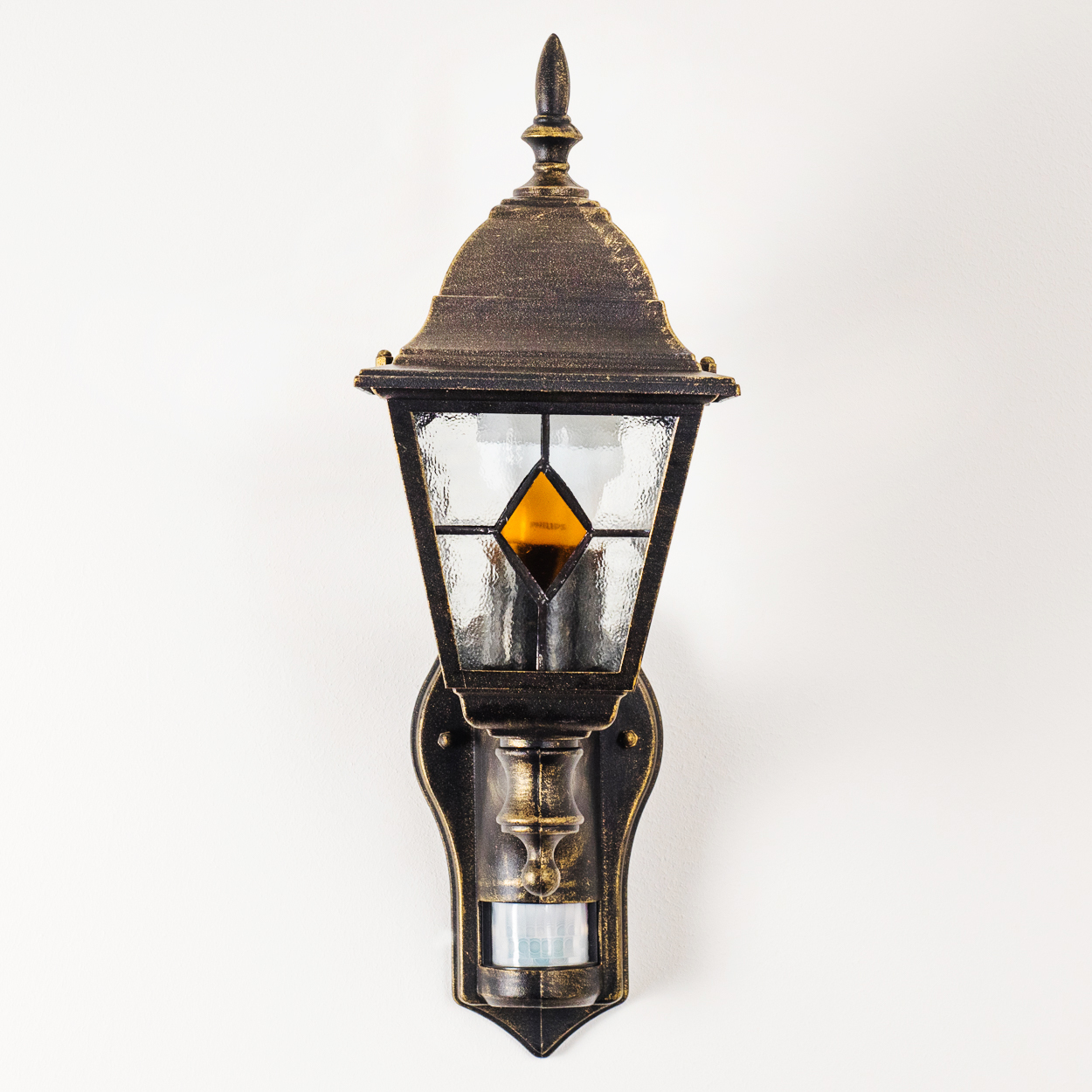 Eine verzierte Außenwandleuchte mit Bronze-Finish. Sie hat eine Glasscheibe mit einem orangefarbenen Diamanten in der Mitte und einen Bewegungssensor.
