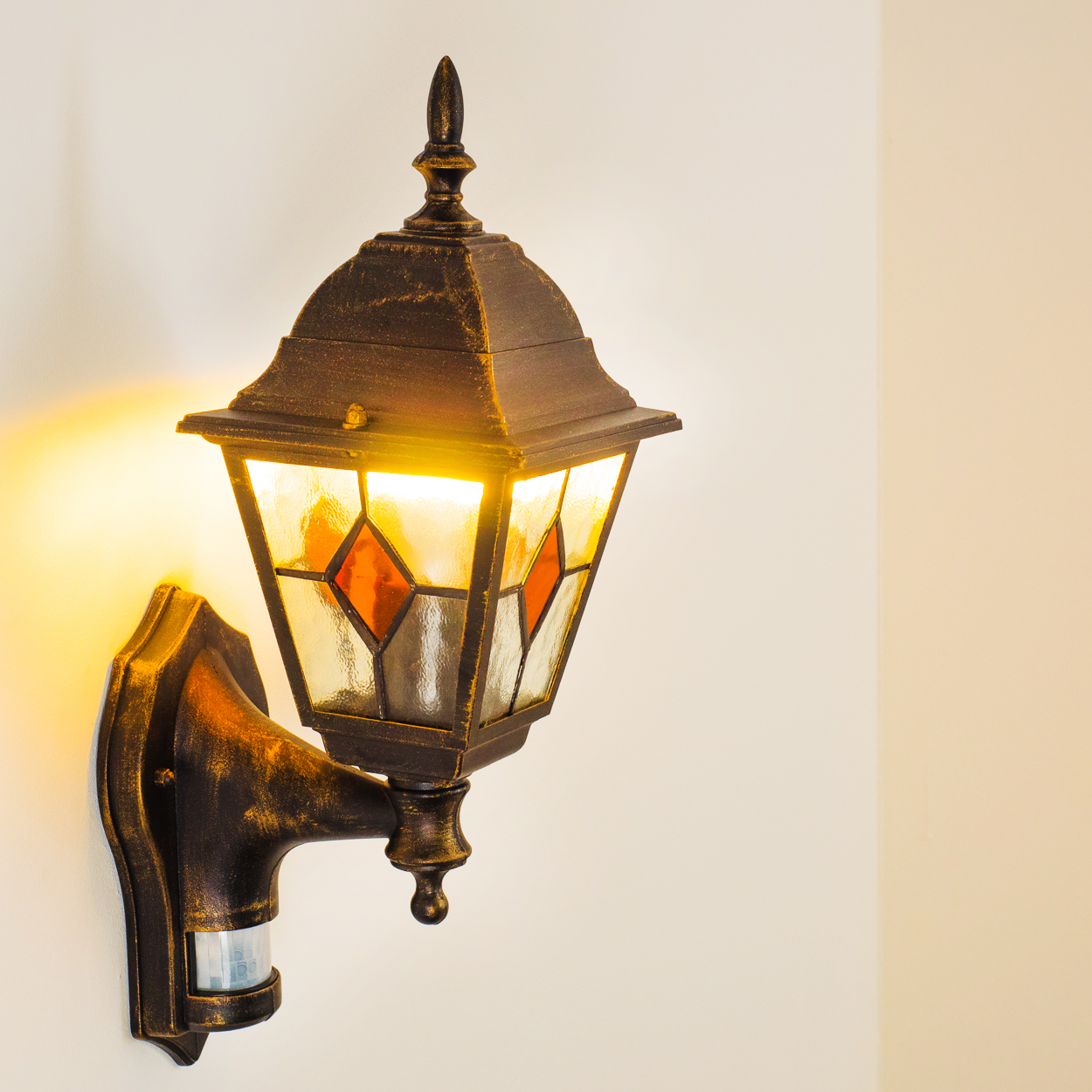 Eine bronzefarbene Außenlampe mit Buntglasscheiben und Bewegungsmelder ist an einer weißen Wand montiert, von der Licht scheint.