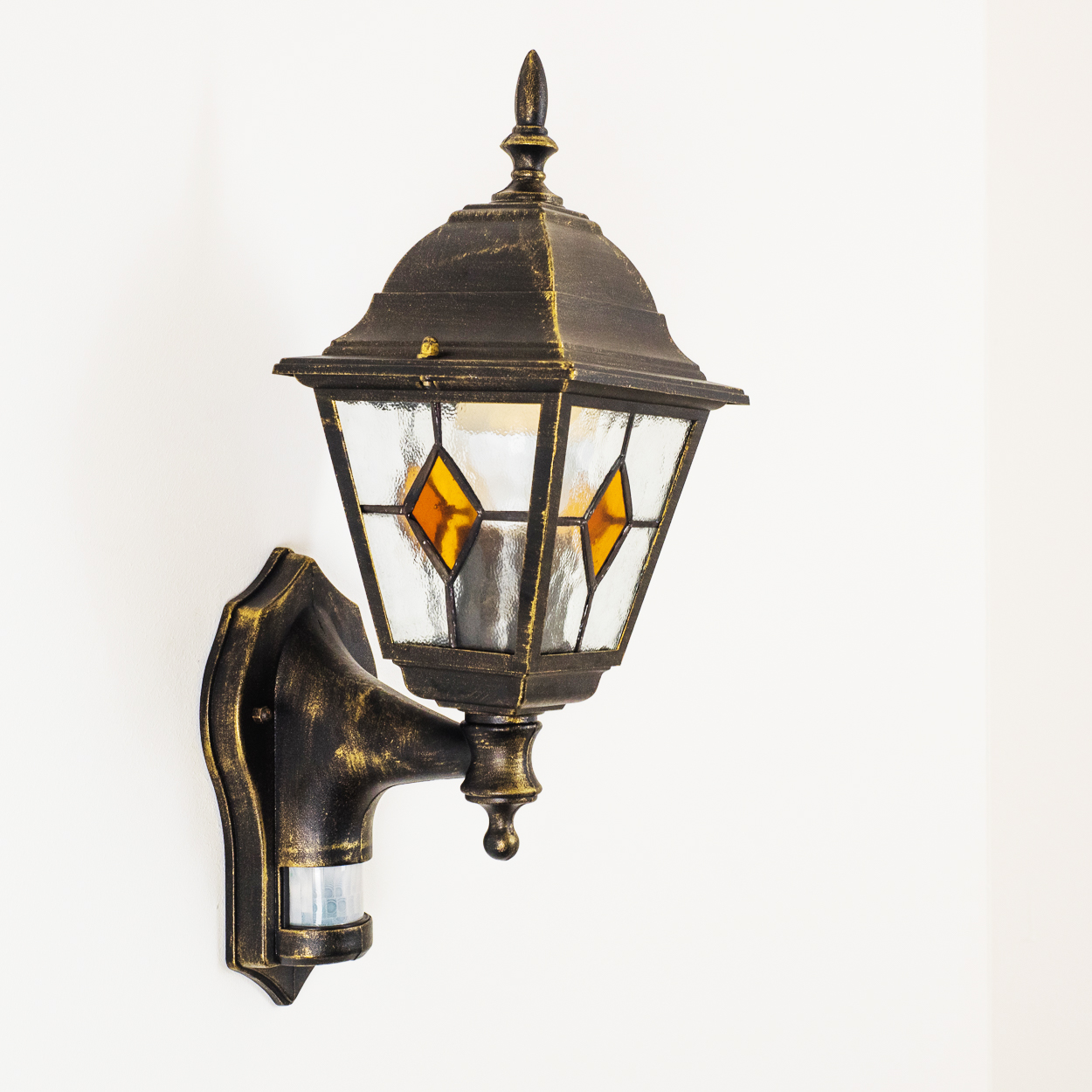 Eine verzierte, bronzefarbene Wandlampe mit Glaspaneelen und gelben Diamantakzenten. Sie ist an einer weißen Wand befestigt.