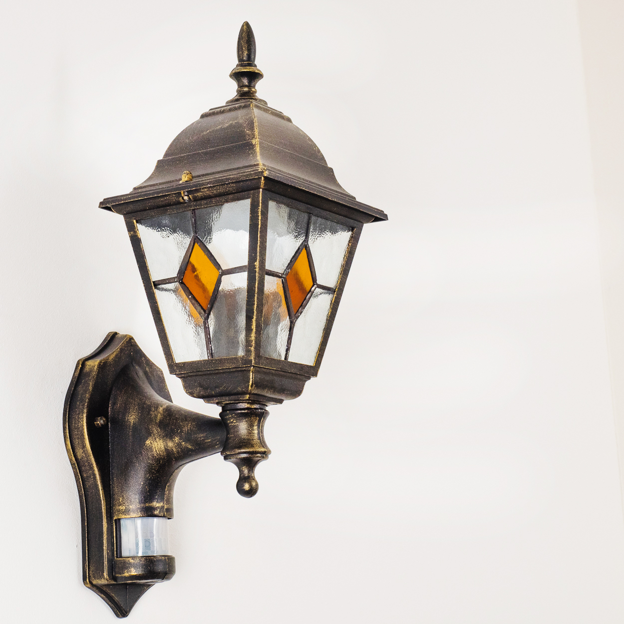 Eine verzierte, bronzefarbene Außenlampe mit Glaspaneelen und einem Bewegungssensor ist an einer weißen Wand montiert.